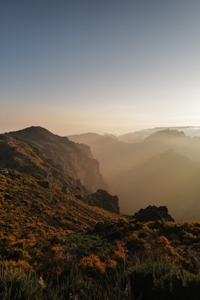 Golden sunlight bathes rolling hills and rugged mountains, casting long shadows over the landscape. The sky transitions from a clear blue at the top to a warm amber near the horizon, with hints of distant clouds.