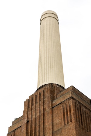 A tall, cylindrical chimney made of white material rising from a large brick building with an industrial architectural style. The chimney and the building both display strong vertical lines, creating a sense of height and angular design.