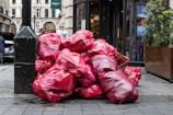 A pile of large red garbage bags is placed against a black pole on a city street. The bags are stacked haphazardly, suggesting an accumulation of waste. In the background, a few people are visible, and there is a storefront with plants flanking the entrance.