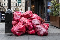 A pile of large red garbage bags is placed against a black pole on a city street. The bags are stacked haphazardly, suggesting an accumulation of waste. In the background, a few people are visible, and there is a storefront with plants flanking the entrance.
