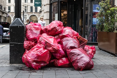 A pile of large red garbage bags is placed against a black pole on a city street. The bags are stacked haphazardly, suggesting an accumulation of waste. In the background, a few people are visible, and there is a storefront with plants flanking the entrance.