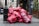 A pile of large red garbage bags is placed against a black pole on a city street. The bags are stacked haphazardly, suggesting an accumulation of waste. In the background, a few people are visible, and there is a storefront with plants flanking the entrance.