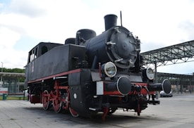 A vintage steam locomotive with a classic black exterior and red wheels is stationary on a paved area. The train features large round headlights and a prominent smokestack, creating a historical and industrial look. In the background, there is a modern urban setting with structures such as metal frameworks and a park-like area.