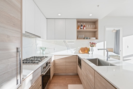 A modern kitchen with light wooden cabinetry and white countertops, featuring a gas stove, sink, and built-in oven. Above the counter are open shelves displaying wine bottles, glasses, and dishes. A small bouquet of flowers and a cutting board add a touch of decoration. Natural light floods the space through a nearby window, creating a bright and airy atmosphere.