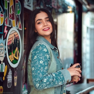 A young woman with long dark hair smiles warmly while holding a paper coffee cup. She is wearing a denim jacket with floral patterns and standing in front of a wall decorated with colorful graffiti art, including a character and vibrant patterns.