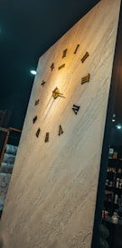 A large wall-mounted clock with Roman numerals, set on a textured, light-colored surface. The clock face is minimalist, with dark hour markers and hands. Surrounding the clock, there are shelves with various items, including boxes and decorative pieces, suggesting an interior setting.