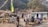People dressed in hiking gear are gathered outside a stone building with a tin roof, likely a rest stop or lodge. There are colorful prayer flags and blooming flowers around the area. The background shows trees and a wooded area.