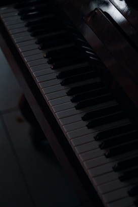 A close-up view of a piano keyboard, highlighting the black and white keys. The lighting is dim, casting shadows and creating a moody atmosphere. The surface of the piano is slightly reflective, adding depth to the image.