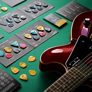 A collection of colorful guitar picks neatly arranged on dark cards, accompanied by a glossy red electric guitar resting on a green surface. The picks vary in shape and design, showcasing a variety of styles.
