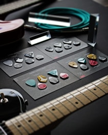 A variety of guitar picks arranged neatly on dark cardstock with labels, placed on a dark surface. Adjacent to the picks are parts of an electric guitar, an audio cable coiled neatly, and what appears to be amplifier knobs. The setup is arranged for display, possibly to showcase the different types of guitar picks available.