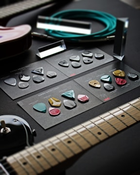 A variety of guitar picks arranged neatly on dark cardstock with labels, placed on a dark surface. Adjacent to the picks are parts of an electric guitar, an audio cable coiled neatly, and what appears to be amplifier knobs. The setup is arranged for display, possibly to showcase the different types of guitar picks available.