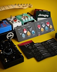 A collection of guitar accessories arranged on a yellow surface, including various guitar pedals, a booklet with guitar picks, and string packages. The devices are colorful, featuring brands like Teisco and Stringjoy, and there is a part of a guitar neck visible in the background.