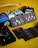 A collection of guitar accessories arranged on a yellow surface, including various guitar pedals, a booklet with guitar picks, and string packages. The devices are colorful, featuring brands like Teisco and Stringjoy, and there is a part of a guitar neck visible in the background.