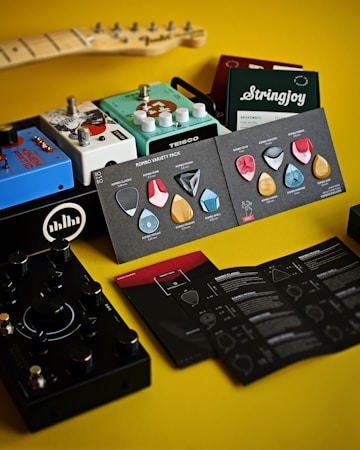 A collection of guitar accessories arranged on a yellow surface, including various guitar pedals, a booklet with guitar picks, and string packages. The devices are colorful, featuring brands like Teisco and Stringjoy, and there is a part of a guitar neck visible in the background.