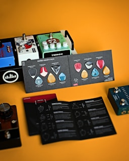A collection of guitar effects pedals sits alongside a product display of guitar picks in varied shapes and colors. The background is a warm orange, complementing the colorful and diverse items, including brand names such as TEISCO and information booklets.
