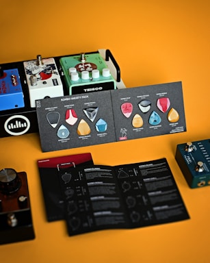 A collection of guitar effects pedals sits alongside a product display of guitar picks in varied shapes and colors. The background is a warm orange, complementing the colorful and diverse items, including brand names such as TEISCO and information booklets.