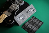 A black electric guitar is displayed next to a variety pack of guitar picks on a green surface. The picks are set on a dark, textured card, and an accompanying booklet is visible with detailed instructions and graphics.