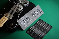 A black electric guitar is displayed next to a variety pack of guitar picks on a green surface. The picks are set on a dark, textured card, and an accompanying booklet is visible with detailed instructions and graphics.