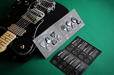 A black electric guitar is displayed next to a variety pack of guitar picks on a green surface. The picks are set on a dark, textured card, and an accompanying booklet is visible with detailed instructions and graphics.