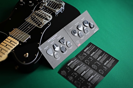A black electric guitar is displayed next to a variety pack of guitar picks on a green surface. The picks are set on a dark, textured card, and an accompanying booklet is visible with detailed instructions and graphics.