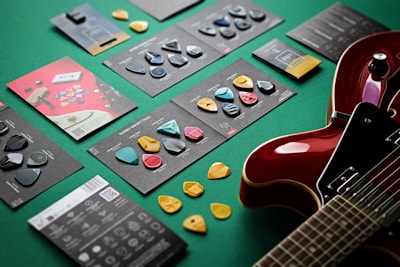 Various guitar picks are displayed on a green surface along with their packaging. A red electric guitar is partially visible on the right side of the image. The picks come in different colors and shapes, and are arranged neatly on black cards with descriptions.