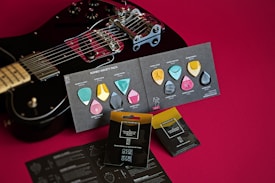 A black electric guitar is placed on a red background alongside a variety of colorful guitar picks displayed on cards. The guitar picks come in multiple shapes and colors, including blue, pink, yellow, and gray. Packaging for a pick holder and other promotional materials are also visible.