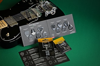 A black electric guitar is placed on a green surface, alongside several packets and cards containing guitar picks. The guitar is positioned diagonally, with chrome hardware and a sleek design. The cards display a variety of picks with different shapes and textures, labeled as a 'Rombo Variety Pack'. Some picks are arranged artistically on the cards. The overall setup suggests a product showcase, balancing the aesthetic appeal of the guitar with the utilitarian design of the picks.