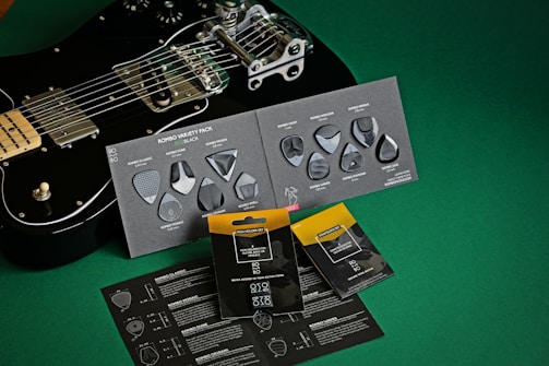 A black electric guitar is placed on a green surface, alongside several packets and cards containing guitar picks. The guitar is positioned diagonally, with chrome hardware and a sleek design. The cards display a variety of picks with different shapes and textures, labeled as a 'Rombo Variety Pack'. Some picks are arranged artistically on the cards. The overall setup suggests a product showcase, balancing the aesthetic appeal of the guitar with the utilitarian design of the picks.