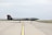 A sleek, dark-colored military drone is positioned on a wide, open runway. The aircraft has a low, elongated shape with minimal markings on its body. The background features a clear sky and patches of grass alongside the runway.