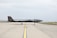 A sleek, dark-colored military drone is positioned on a wide, open runway. The aircraft has a low, elongated shape with minimal markings on its body. The background features a clear sky and patches of grass alongside the runway.