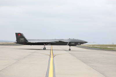 A sleek, dark-colored military drone is positioned on a wide, open runway. The aircraft has a low, elongated shape with minimal markings on its body. The background features a clear sky and patches of grass alongside the runway.
