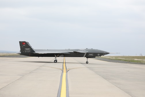A sleek, dark-colored military drone is positioned on a wide, open runway. The aircraft has a low, elongated shape with minimal markings on its body. The background features a clear sky and patches of grass alongside the runway.