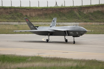 A sleek, modern military aircraft is positioned on a runway. The aircraft is angular in design, featuring a dark grey coloration with various markings and insignia. The background includes a grassy area and a fence line, suggesting an airfield setting.