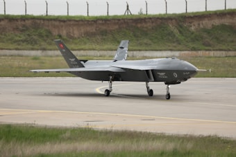 A sleek, modern military aircraft is positioned on a runway. The aircraft is angular in design, featuring a dark grey coloration with various markings and insignia. The background includes a grassy area and a fence line, suggesting an airfield setting.