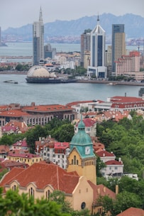 An urban landscape featuring a waterfront cityscape with modern skyscrapers and a distinctive building with a clock tower in the foreground. Red-roofed buildings and lush greenery add contrast, while distant mountains are visible in the background.