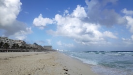 A sandy beach stretches along the coastline with gentle waves lapping against the shore. On the left, a line of resort buildings with palm trees are visible, extending into the distance. The sky is mostly clear with large, fluffy clouds scattered across a bright blue backdrop.