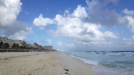 A sandy beach stretches along the coastline with gentle waves lapping against the shore. On the left, a line of resort buildings with palm trees are visible, extending into the distance. The sky is mostly clear with large, fluffy clouds scattered across a bright blue backdrop.