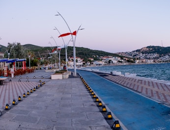A seaside promenade or walkway with distinct blue and stone-paved paths. Red flags wave atop modern streetlights lining the path. To the left, several outdoor seating areas belong to waterfront cafes or restaurants. The area opens up to a view of the sea, with waves gently reaching the shore. In the background, a hilly residential area with tightly packed buildings stretches along the coastline.