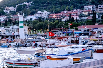 A bustling marina filled with multiple boats docked along the waterfront. Many of the boats are adorned with red flags featuring a white star and crescent. In the background, there is a mountainous area with numerous trees and residential buildings with colorful roofs. A prominent white and green striped tower is visible near the dock.