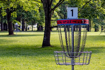 In a spacious park setting, a disc golf target with chains and a basket is prominently displayed in the foreground, marked with the number one. Lush green grass and tall trees surround the area, offering a serene and natural backdrop. A second disc golf basket is visible in the distance.