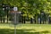 A disc golf basket is situated in a lush green park setting, surrounded by trees with bright green leaves. The basket is marked with a number '6', and chains are visible hanging from the top of the basket structure. Sunlight filters through the leaves, casting dappled shadows on the grass.