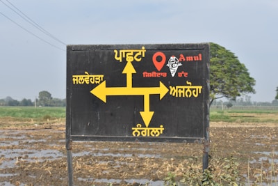 A black signboard with yellow directional arrows and text written in Punjabi, indicating directions to various locations. The sign includes the word 'Amul' and an image of an ice cream cone. The background shows a rural landscape with a field and some trees.