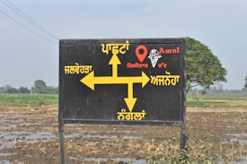 A black signboard with yellow directional arrows and text written in Punjabi, indicating directions to various locations. The sign includes the word 'Amul' and an image of an ice cream cone. The background shows a rural landscape with a field and some trees.