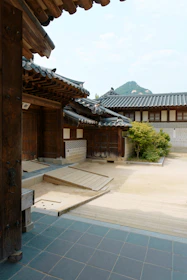 A traditional wooden Korean architecture with tiled roofs and a courtyard featuring stone flooring and wooden decks. A small garden with a bush is present, and a mountain is visible in the background against a partly cloudy sky.