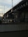 A large bridge structure dominates the view, with visible pillars and support beams. Overhead electrical lines and poles are present, suggesting it might be a railway bridge. Below, a construction area with fences, equipment, and a vehicle can be seen. The scene is partially obscured by shadows, with sunlight illuminating parts of the structure.