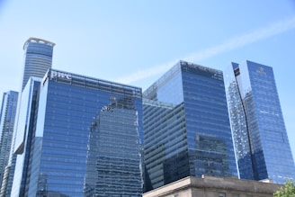 A group of modern skyscrapers with reflective glass facades, showcasing a blend of contemporary architecture. Some buildings have prominent logos visible on their surfaces.