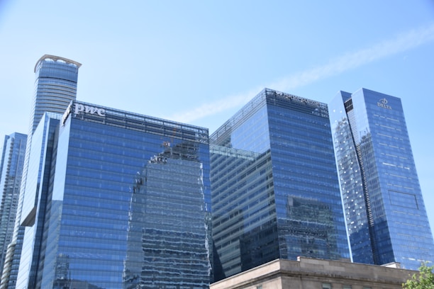 A group of modern skyscrapers with reflective glass facades, showcasing a blend of contemporary architecture. Some buildings have prominent logos visible on their surfaces.
