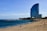 A tall, modern hotel with a distinctive curved design stands near a sandy beach. The ocean is calm, with gentle waves running parallel to the shoreline. Several people are relaxing on the beach, some walking close to the water, while a breakwater of rocks extends into the sea. Palm trees are visible near the hotel, adding to the coastal atmosphere.