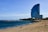 A tall, modern hotel with a distinctive curved design stands near a sandy beach. The ocean is calm, with gentle waves running parallel to the shoreline. Several people are relaxing on the beach, some walking close to the water, while a breakwater of rocks extends into the sea. Palm trees are visible near the hotel, adding to the coastal atmosphere.