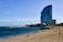 A tall, modern hotel with a distinctive curved design stands near a sandy beach. The ocean is calm, with gentle waves running parallel to the shoreline. Several people are relaxing on the beach, some walking close to the water, while a breakwater of rocks extends into the sea. Palm trees are visible near the hotel, adding to the coastal atmosphere.